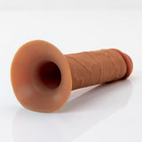 Alt text: A FX Elite 6 Silicone Hollow Strap-On in tan, with a dual-density design and textured skin-like exterior, rests on a white surface. It features a circular opening at one end and a rounded tip at the other.