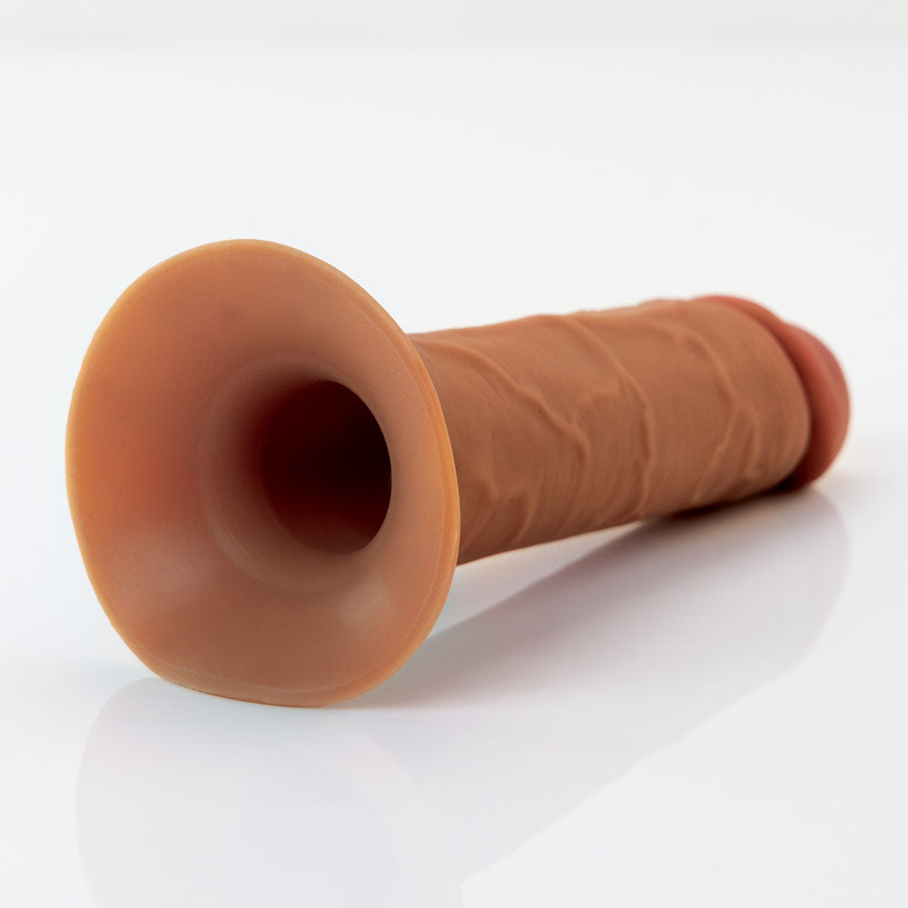 An FX Elite 7 Silicone Hollow Strap-On in tan, featuring a realistic design with a textured, veined surface and rounded tip, is laid flat against a white background. Its dual-density structure and strong suction cup base ensure hands-free stability.