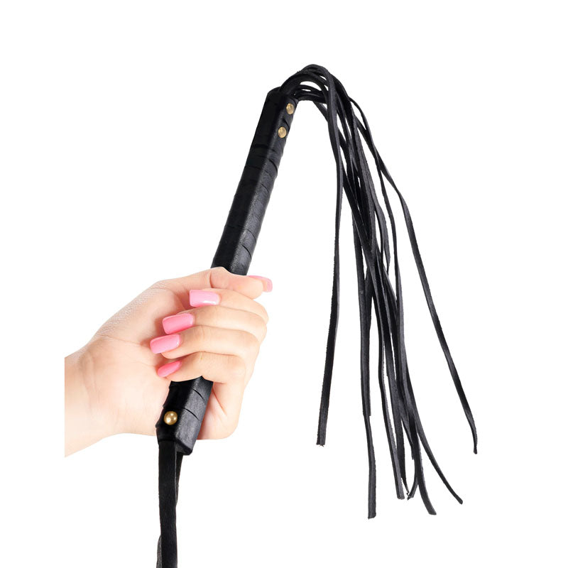 A hand with pink manicured nails holds the Fetish Fantasy Series Limited Edition Black Cat-o-nine Tails whip, featuring a braided handle with gold studs and multiple leather tails. The scene is set against a plain white background, inviting you to play hard.