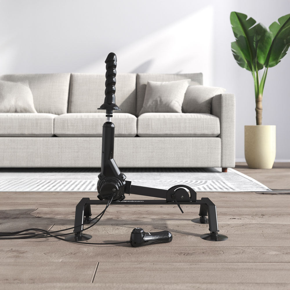 A sleek black FFS Body Dock Sex Machine is positioned on a wooden floor in front of a grey sofa with pillows. A large potted plant on the right complements the bright and minimalistic room, which features a white and grey area rug under the sofa.