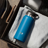 Inside an open dresser drawer, a blue, insulated PDX Plus Fuck Flask is placed on a light gray t-shirt. A folded white towel and stacks of dark clothes are in the background, with the drawer handle visible on the left.