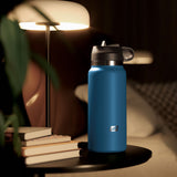 A blue, insulated water bottle with a black lid sits discreetly on a round black table beside a stack of books. A modern lamp with a white shade illuminates the scene, enhancing its charm. In the blurred background are a cushioned chair and patterned pillow. Product: PDX Plus Fuck Flask - Private Pleaser - Flesh Discreet Vagina Stroker.