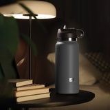 A black insulated PDX Plus Fuck Flask - Secret Delight sits discreetly on a round, dark table. Nearby are stacked books and a modern lamp with a warm glow. A blurred plant and checkered pillow create a cozy atmosphere, concealing its dual purpose as an innovative water bottle stroker.