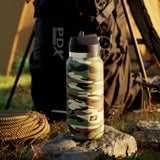 The PDX Plus Fap Flask - Happy Camper, a frosted discreet stroker in camo pattern, sits on a rock with coiled rope. A black backpack and beige tent stand in the background, creating a camping scene. Sunlight casts warm tones, enhancing the outdoor atmosphere.