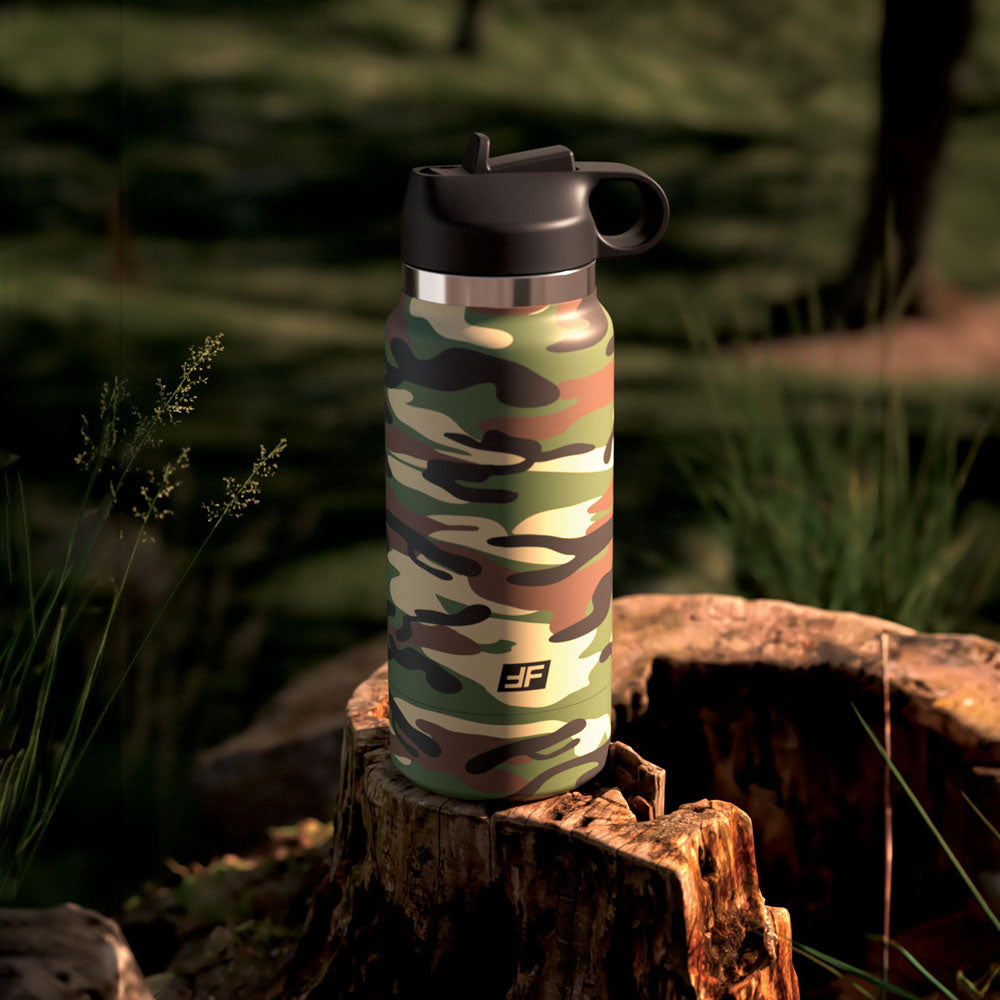 A PDX Plus Fap Flask - Happy Camper sits on a tree stump in a sunlit forest. Grass and blurred foliage surround it, casting soft shadows. The initials JF are on the bottle, blending with the natural backdrop.