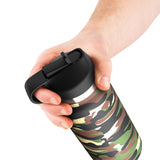 A hand holding a camo-patterned PDX Plus Fap Flask - Happy Camper with a black lid and loop handle, featuring green, brown, and beige camouflage on a plain white background.