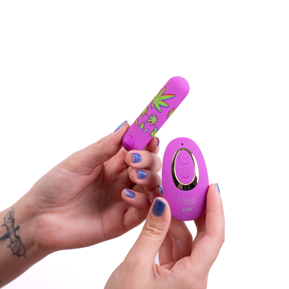 Two hands hold a Maia JESSI 420 Remote - Purple 7.6 cm Bullet with a silicone coating and matching wireless remote. The person, with purple nail polish and a bat tattoo on their wrist, showcases the submersible design against a white background.