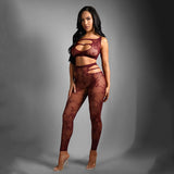 A woman poses gracefully against a gray background in the SHEER FANTASY UNDIVIDED ATTENTION bodysuit, featuring burgundy patterns with rhinestone accents and cutouts. Her long dark hair cascades elegantly as she wears clear high heels, her right hand on her thigh and left on her hip.