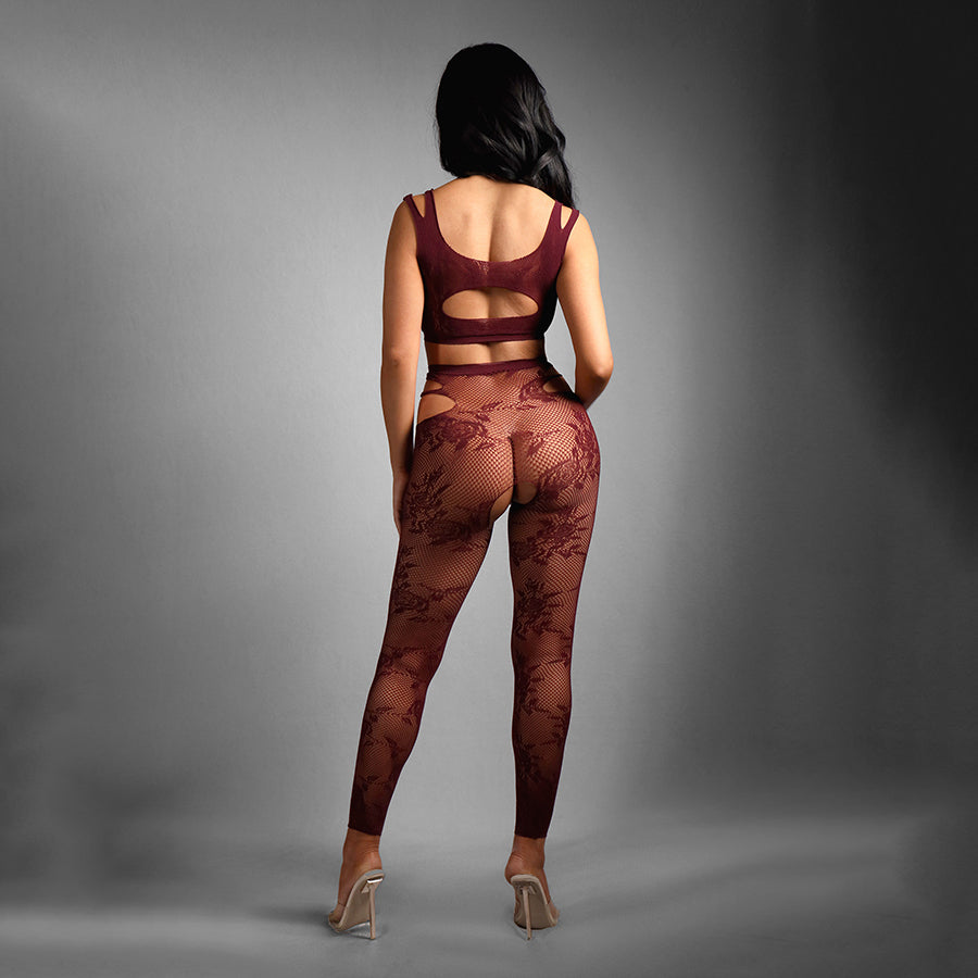 A person with long dark hair stands against a gray background, facing away from the camera, wearing the SHEER FANTASY UNDIVIDED ATTENTION set in burgundy. The top has cut-out details and is paired with matching floral-patterned footless tights and clear high-heeled shoes.