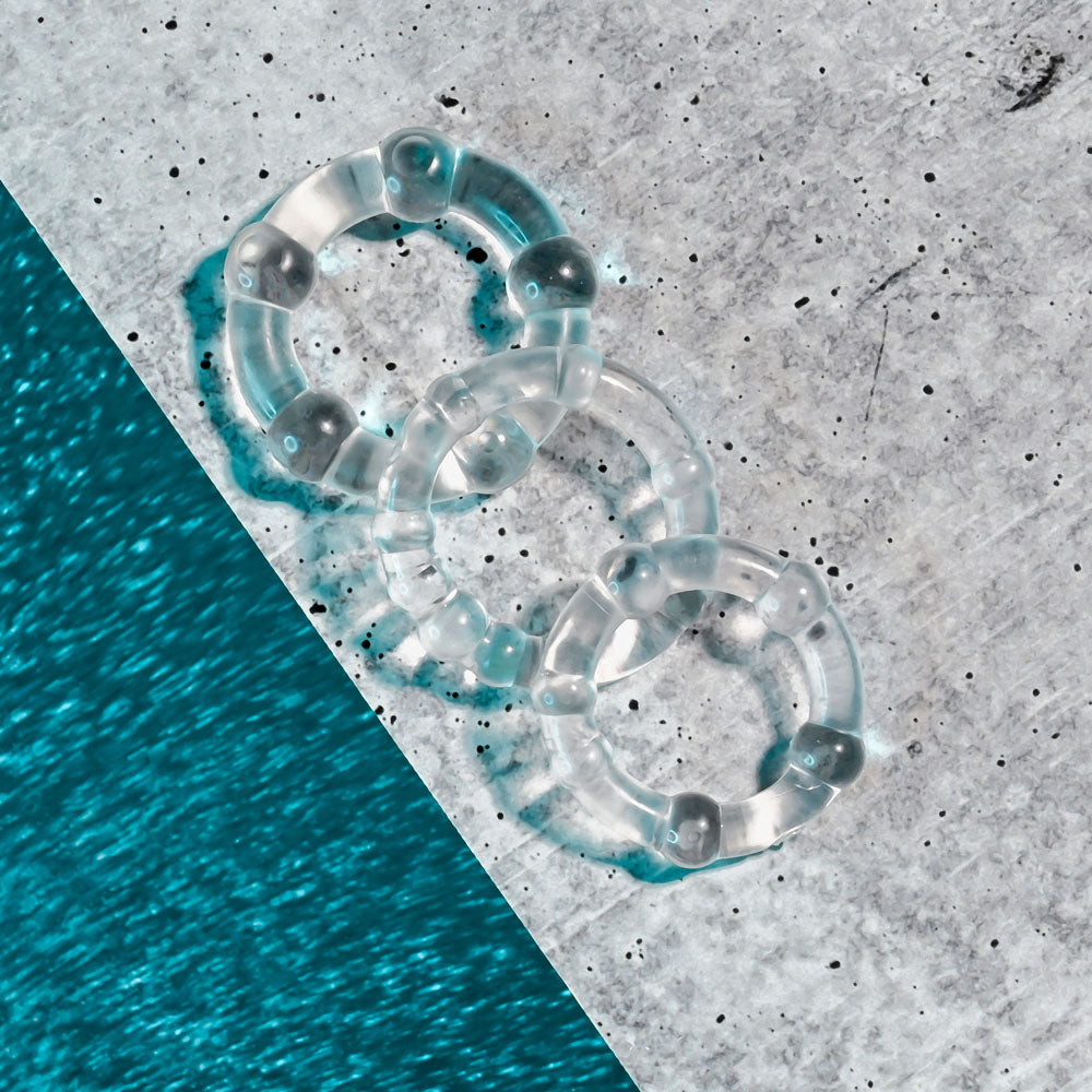 The Selopa ERECTION RINGS - Clear Cock Rings Set of 3, featuring smooth, glossy interconnected rings, rest on a textured speckled background. A blue wavy pattern diagonally divides the scene, highlighting reflections on the super-stretchy rings.