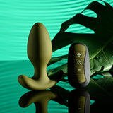 The Selopa THE COLONEL, a green 10.1 cm USB rechargeable vibrating butt plug with a flared base, and its black wireless remote featuring plus, minus, and power buttons rest on a reflective surface with a large green leaf in the background, creating a serene and modern ambiance with soft shadows.