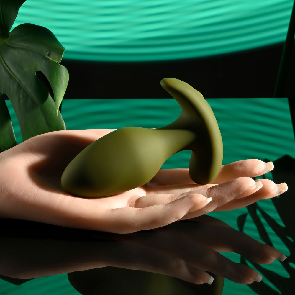 A close-up shows Selopa THE COLONEL, a green phthalate and latex-free silicone anal plug, on a mannequins hand against a reflective surface. A large tropical leaf and striped teal pattern in the background highlight the plugs smooth matte finish.