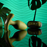 The Selopa THE COLONEL, a green 10.1 cm vibrating silicone butt plug, is paired with a wireless remote featuring plus, minus, and power buttons. It rests on a reflective surface against monstera leaf silhouettes and a wavy green backdrop for a tropical vibe.