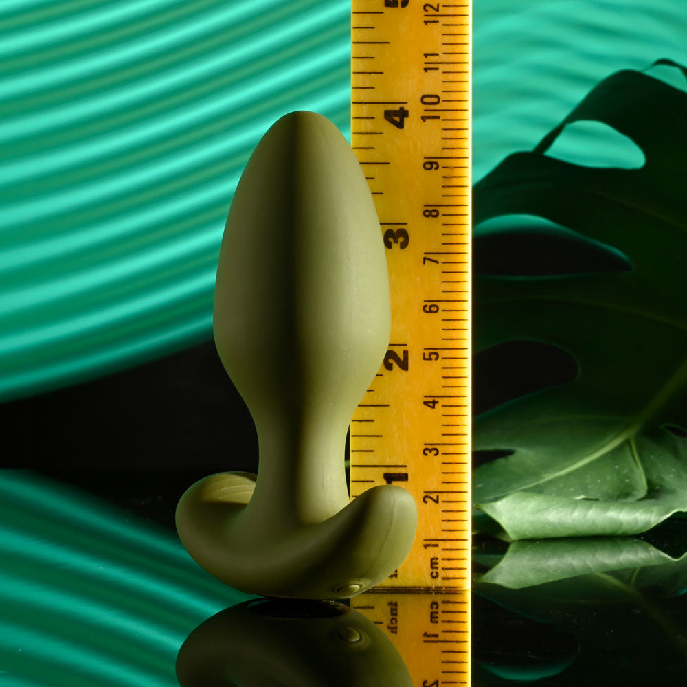 The Selopa THE COLONEL, a sleek green 10.1 cm USB rechargeable vibrating butt plug, stands upright against a yellow ruler with a large leaf and wavy fabric in the background. A reflective surface mirrors the plug and part of the ruler, enhancing its elegant, earthy ambiance.