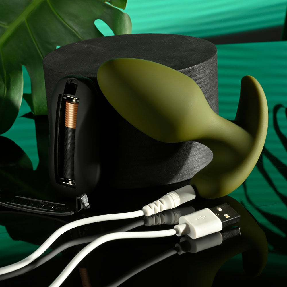 Displayed is the Selopa THE COLONEL, a 10.1 cm green teardrop-shaped USB rechargeable silicone butt plug with a wireless remote, next to a black cylindrical case with visible copper coils. Two white multi-connector USB cables rest on a reflective surface against a striped green backdrop with a large leaf.