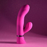 The Selopa NEXT WAVE - Pink 16.4 cm USB Rechargeable Dual Vibrator stands upright against a glittery pink background, featuring a smooth and ribbed texture, dual prongs with a main shaft and smaller curved extension, powerful motors, and a metallic ring near the base for grip or adjustment.