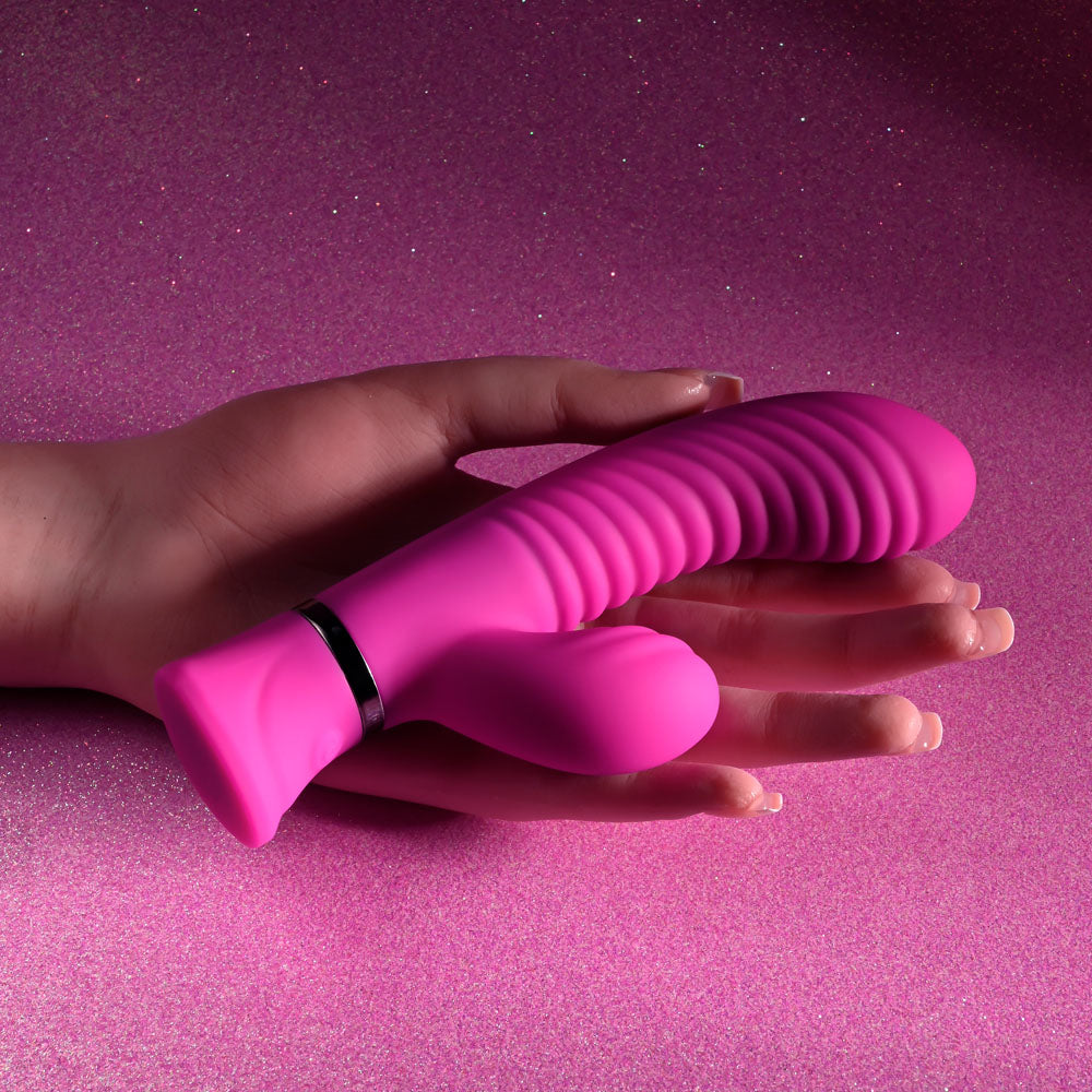 A hand holds the Selopa NEXT WAVE - Pink 16.4 cm USB Rechargeable Dual Vibrator with powerful motors against a glittery pink background. The device has a ribbed, curved design, featuring a metallic ring accent and a small attachment at the base.