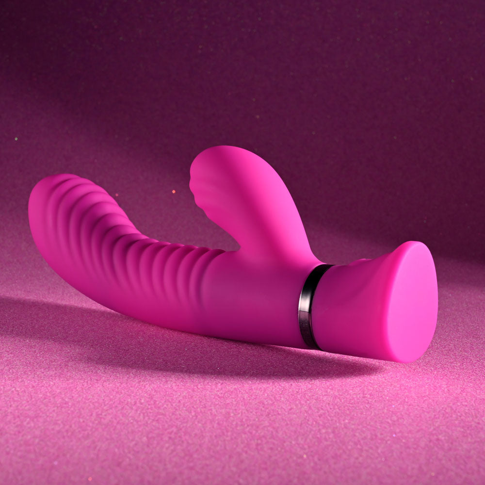 The Selopa NEXT WAVE - Pink 16.4 cm USB Rechargeable Dual Vibrator, with its pink silicone body and ribbed shaft, features a smaller clitoral stimulator and powerful motors. A metallic ring adorns its base as it rests on a textured pink surface, while the lighting casts a soft, colorful ambiance.