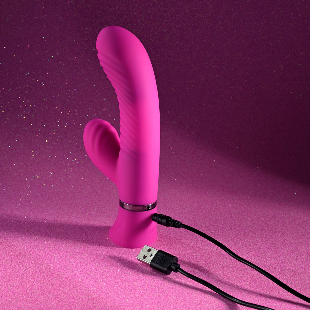 The Selopa NEXT WAVE - Pink 16.4 cm USB Rechargeable Dual Vibrator, featuring a ribbed design and curved shape with a secondary arm for dual stimulation, stands on a glittery pink surface connected to a black USB cable. Its vibrant color is beautifully complemented by the shimmering background.