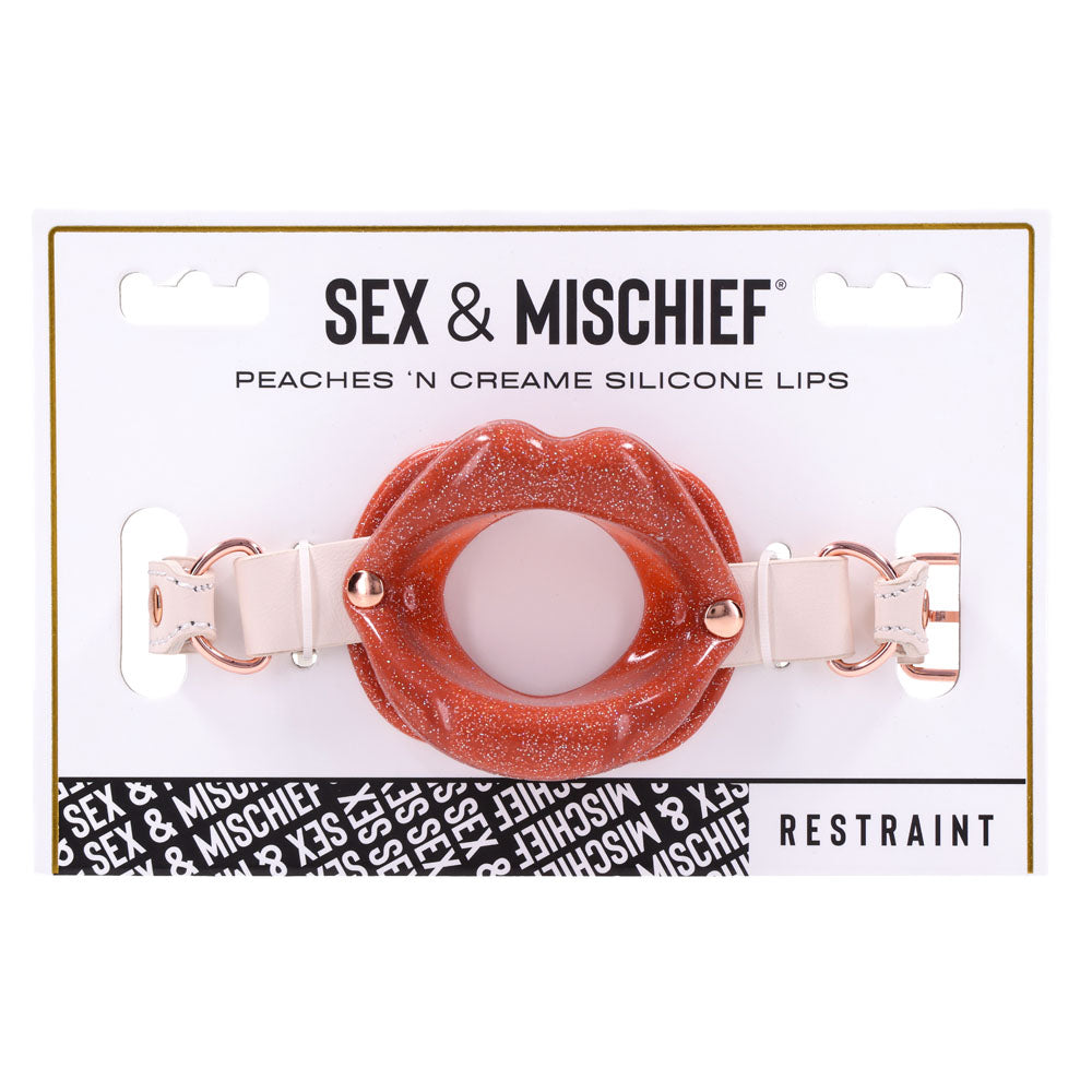 The Sex & Mischief Peaches n CreaMe Silicone Lips packaging showcases a glossy red silicone lip gag with an open center, secured by adjustable faux leather straps. The word Restraint is at the bottom right, with a black and white pattern strip on the left.