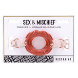 The Sex & Mischief Peaches n CreaMe Silicone Lips packaging showcases a glossy red silicone lip gag with an open center, secured by adjustable faux leather straps. The word Restraint is at the bottom right, with a black and white pattern strip on the left.