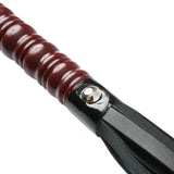 A close-up of the Sex & Mischief Mahogany Flogger displays its black tail and shiny, ribbed dark red handle with wood accents. Nickel-free metal fastenings connect the handle to the tail, all set diagonally on a white background.