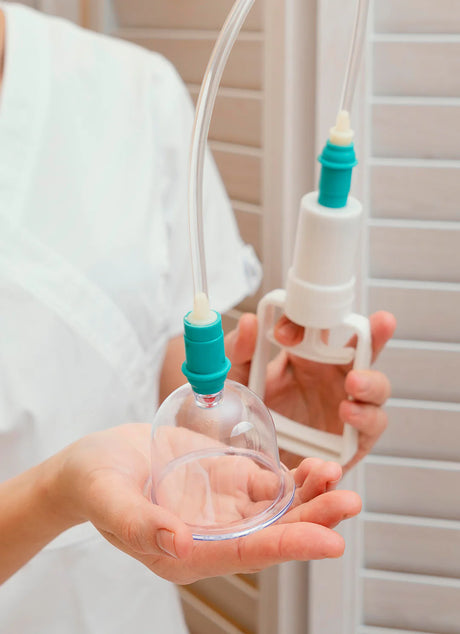 A person holding a clear breast pump with a green and white handle. The pump has a transparent cup and two tubes extending from it. The background features vertical white blinds. The persons arm is visible, wearing a white short-sleeve garment.