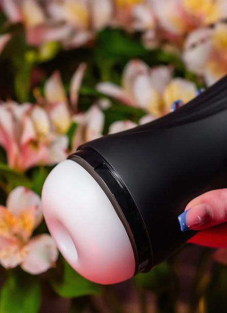 A close-up of a persons hand holding a sleek black cylinder-shaped device with a rounded white end. The background features soft-focus pink and white flowers with green leaves, adding a contrasting natural element to the scene.