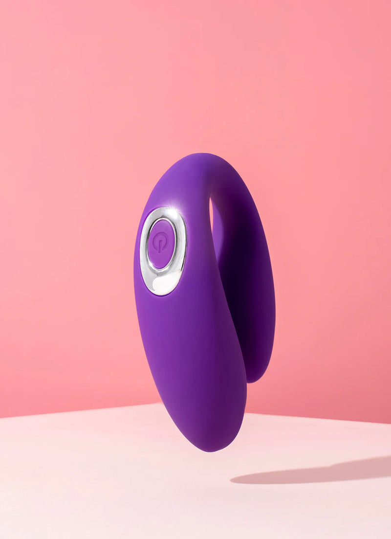 A purple, U-shaped personal massager floats against a dual-toned pink background. It has a circular control button with a power icon in the center. The objects sleek design features smooth curves and a glossy finish, casting a soft shadow on the surface below.