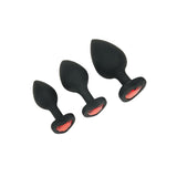 The WhipSmart Heartbreaker 3PC Silicone Crystal Heart Anal Training Set consists of three black butt plugs with red heart gem bases, displayed diagonally from smallest to largest. Ideal for body exploration, their smooth and tapered design elegantly contrasts against a plain white background.