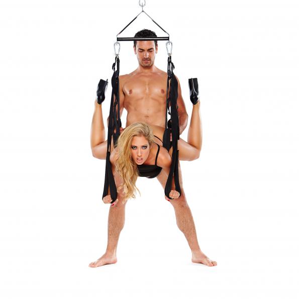 A couple is engaged in a suspended pose using the WhipSmart Pleasure Swing - Black Adjustable Swing. The man stands behind holding the straps, while the woman, dressed in black, is upside down with bent legs over her shoulders against a plain white background, creating an intimate ambiance.