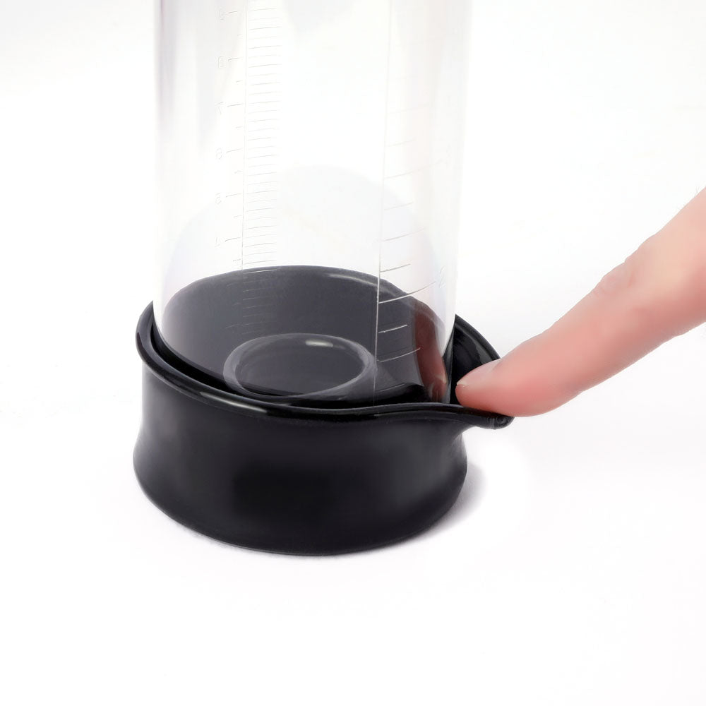 The LuvPump ProXtra is a clear cylindrical penis pump with measurement markings, standing vertically. It has a black rubber base gently pressed by an index finger, slightly deformed, against a plain white background.