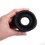 A hand holds a black, doughnut-shaped silicone object from the LuvPump ClassiXtra Advanced Power Pump, resembling a penis pump component. The smooth, rounded gadget is shown against a plain white background.