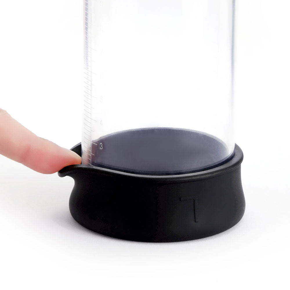 A finger presses the black silicone cover at the base of a clear cylindrical LuvPump Smart Penis Pump, which features side measurement markings. The flexible indented base and vacuum stimulation design enhance performance, making it an innovative choice. USB rechargeable for convenience.