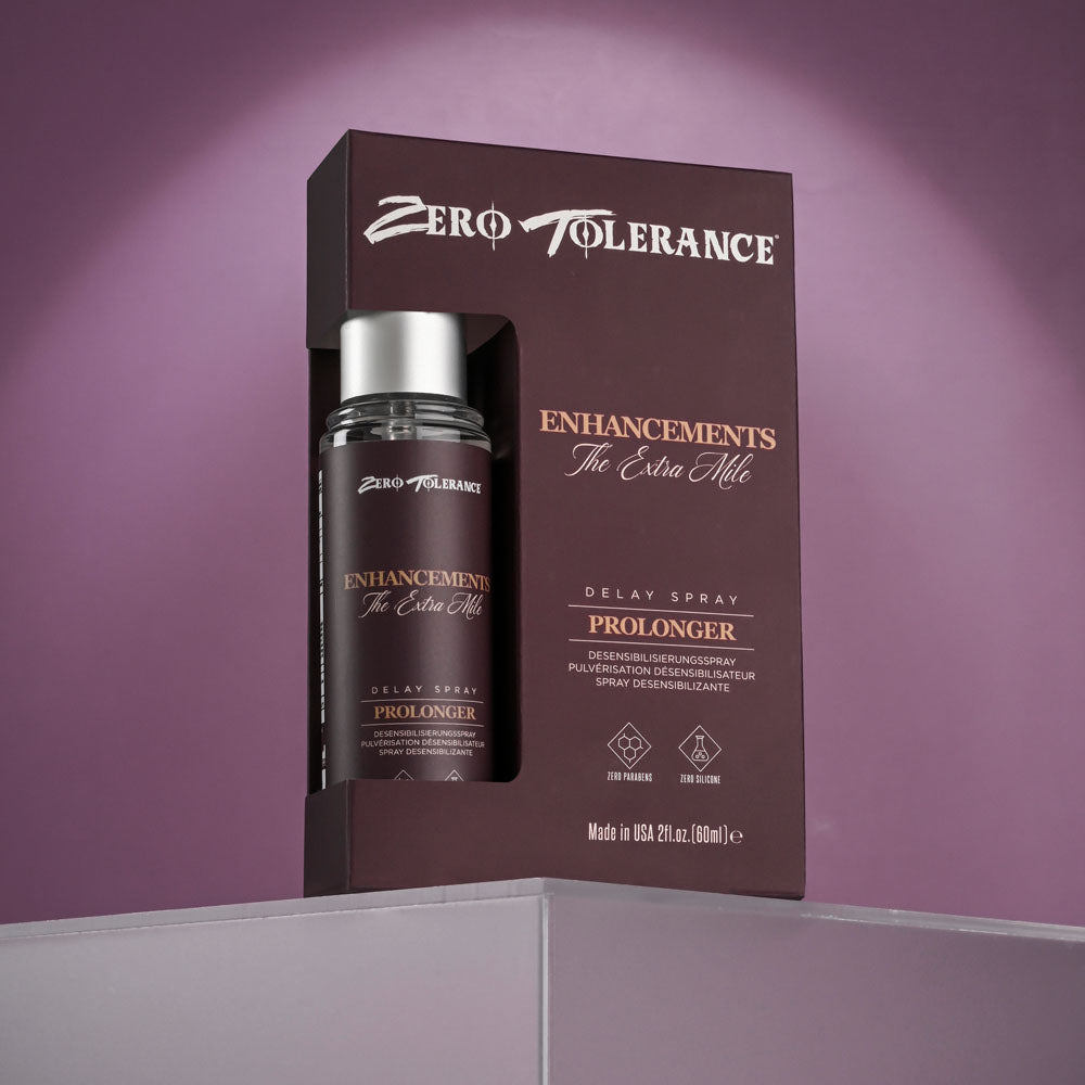 A dark brown box with silver text displaying Zero Tolerance Enhancements - The Extra Mile delay spray for men (60 ml) is partially open, revealing the bottle inside. It sits on a gray surface against a soft purple background.