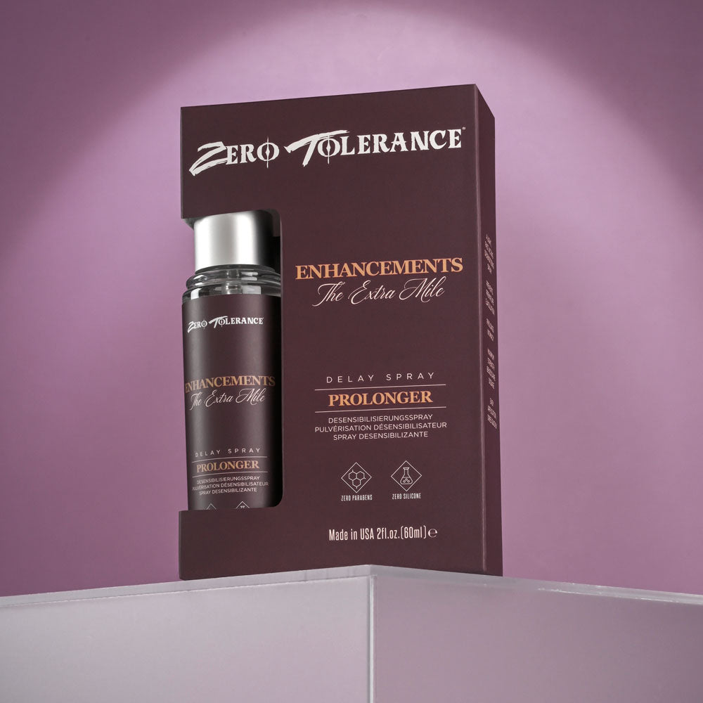 The image shows a bottle of Zero Tolerance Enhancements - The Extra Mile - Delay Spray for Men (60 ml) next to its metallic-lettered dark purple box on a reflective white surface, with a purple background, promising enhanced performance and stamina.