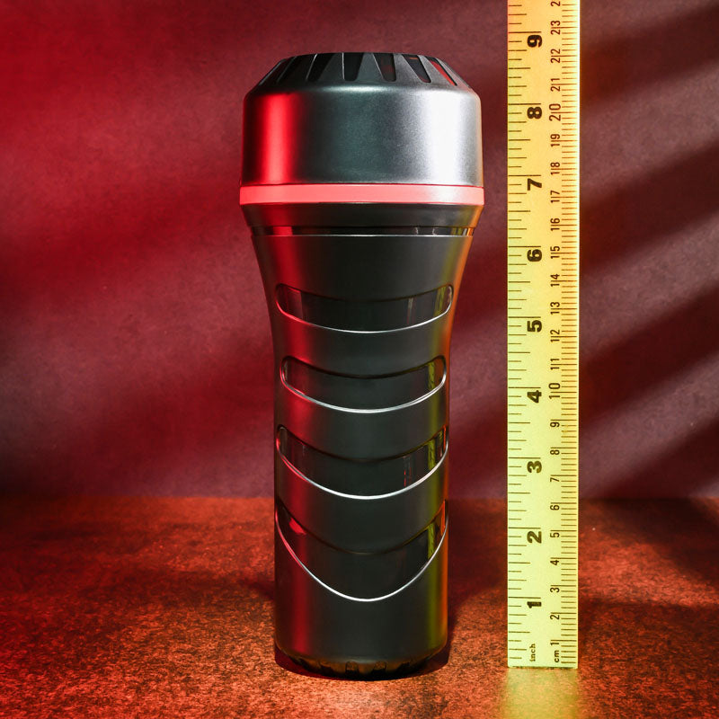 The Zero Tolerance POP ON THE GO Dark - Brown Vagina Masturbator, with a textured grip, stands upright beside a yellow measuring ruler. Red and green lighting casts shadows as the ruler indicates its about 9 inches tall against a dark, textured background.