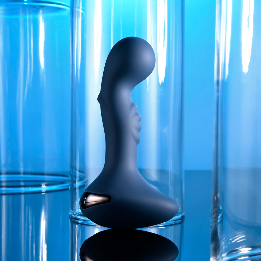 The Zero Tolerance BLUE OASIS, a dark blue, curved 12.3 cm vibrating anal plug with ribbed details and ergonomic grip, stands upright in a clear cylindrical glass container. The glossy background and reflections create a futuristic, vibrant blue ambiance.