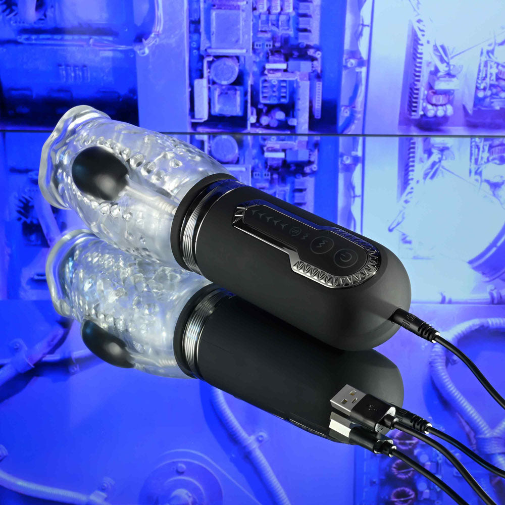 A Zero Tolerance THRILL RIDE in black and clear, featuring a textured cylinder, sits on a reflective surface with two USB cables. The blue-toned background of machinery and electronics adds to the tech-inspired aesthetic of this thrusting, rotating, and vibrating stroker.