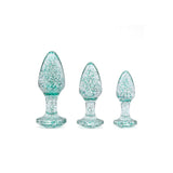 The Adam & Eve Glitter Gem 3-Piece Butt Plug Set features three clear glass plugs of increasing size, each filled with teal glitter. They have a smooth, bulbous shape with a tapered tip and a flared base for stability against the white background.