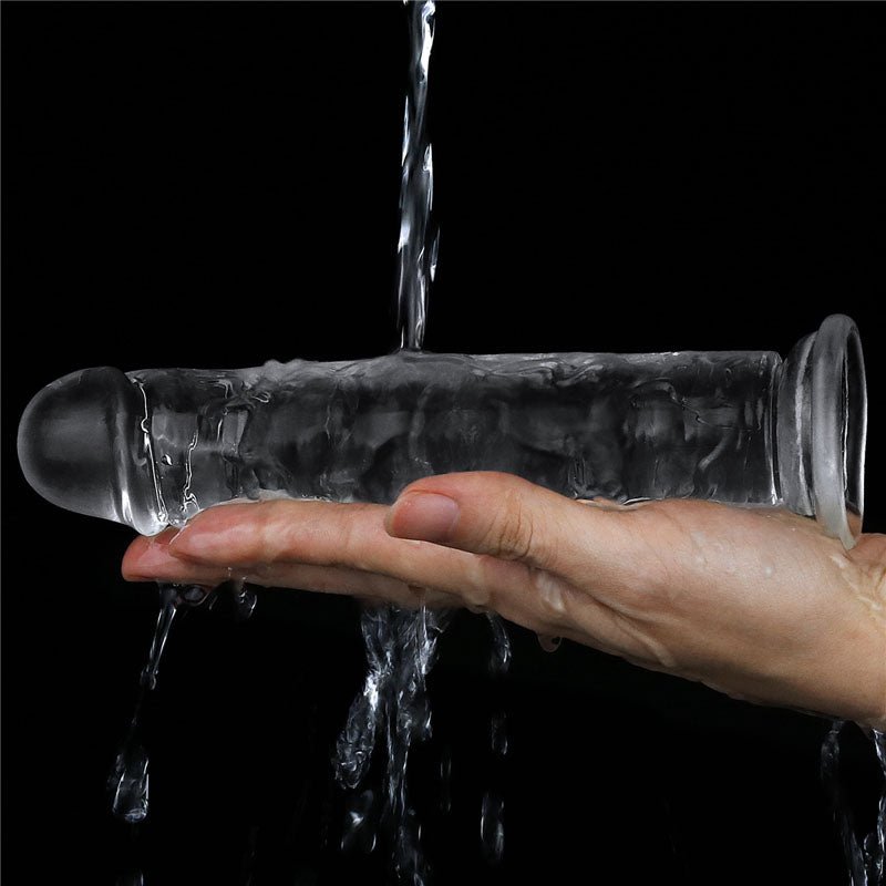 A hand holds a LOVETOY Flawless Clear Dildo 7 - Clear 17.8 cm Dong with rounded ends, as water cascades over the skin-safe transparent toy, creating a flowing effect against a black background.